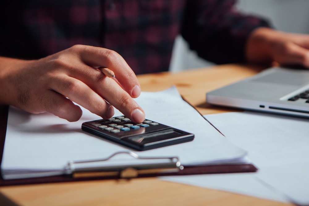 Uma mão a utilizar uma calculadora sobre documentos, com um portátil desfocado ao fundo.