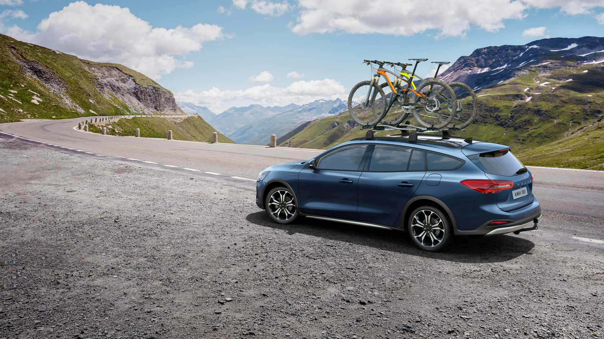Ford Focus azul com bicicletas no tejadilho a percorrer uma paisagem de estrada montanhosa.
