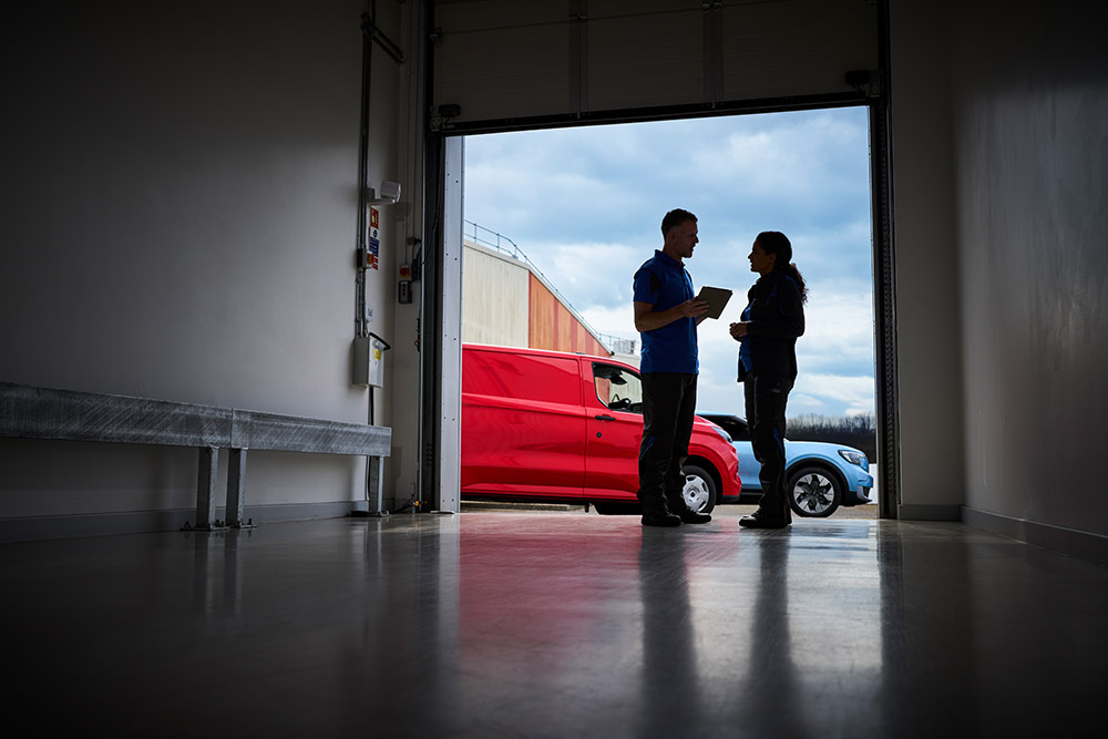 Duas pessoas a conversar à porta de uma garagem, com uma carrinha Ford vermelha e um SUV Ford azul estacionados no lado de fora.