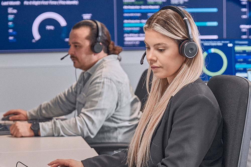  Duas pessoas com auscultadores a trabalhar em computadores numa sala de controlo moderna.