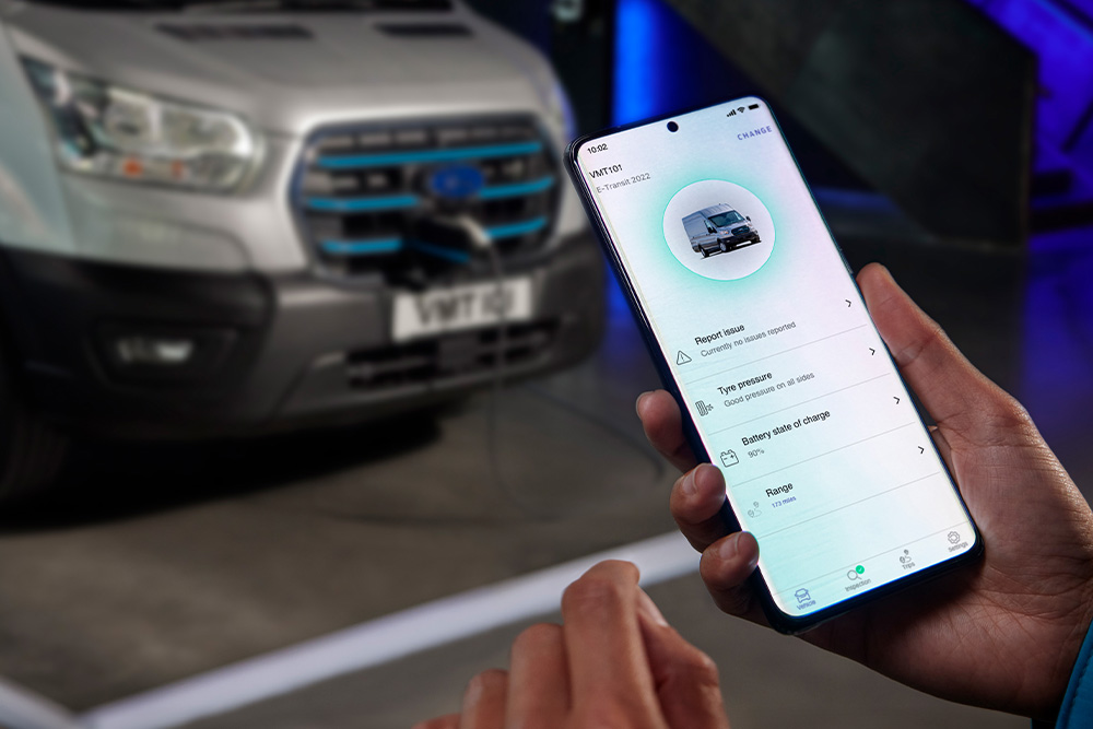 Uma pessoa segura um smartphone com uma aplicação Ford, com um Ford E-Transit Custom a carregar no fundo.