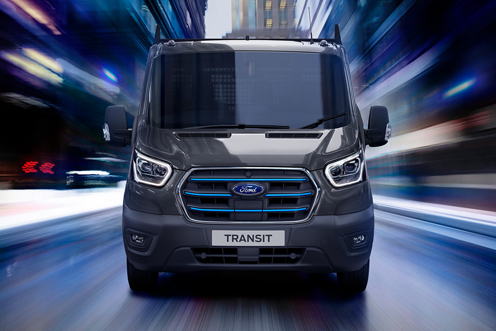 Vista frontal de uma carrinha Ford Transit cinzento-escura em movimento, com um fundo desfocado.
