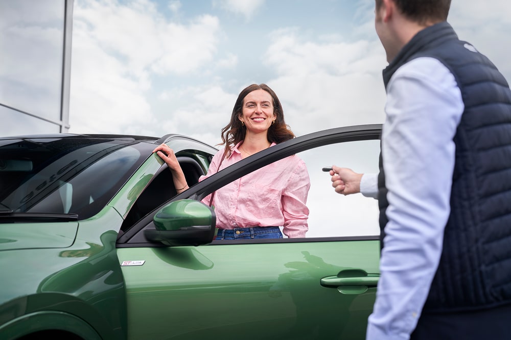 Mulher com camisa cor-de-rosa a sorrir ao lado de um carro verde, com alguém de um concessionário Ford a entregar-lhe as chaves do carro.