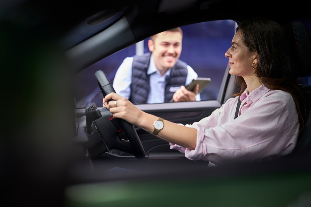 Mulher num carro verde estacionado a sorrir para alguém de um concessionário Ford do lado de fora da janela.